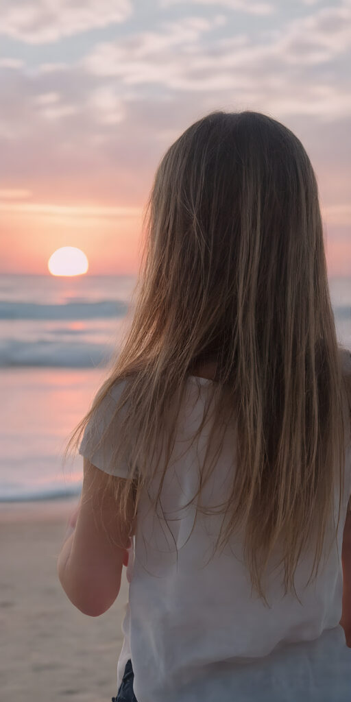 a girl stands on a lonely beach, she enjoy a beautiful sunset and the breaking waves in the background. She has very long straight soft hair, which falls over her upper body, view from the back